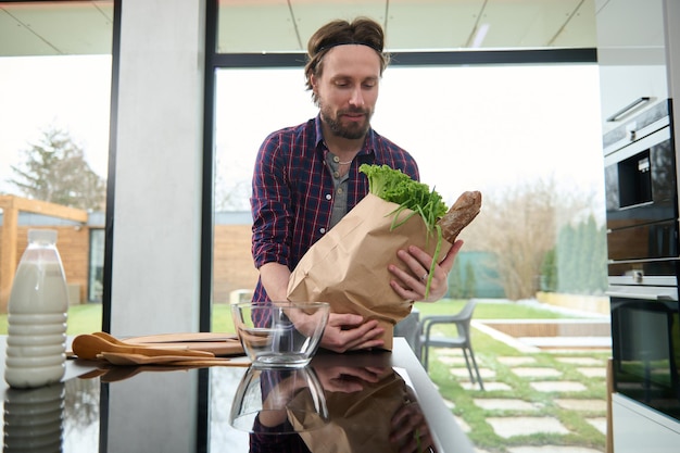 Handsome Caucasian guy, charming young happy man puts ecological paper bag with healthy food on the kitchen table and stands near the large windows overlooking the garden in a spacious villa