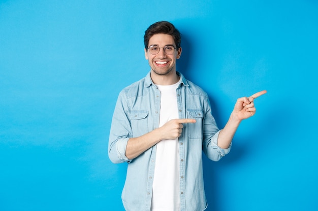 Handsome caucasian male model in glasses, pointing fingers right at your logo, showing copy space, standing over blue background