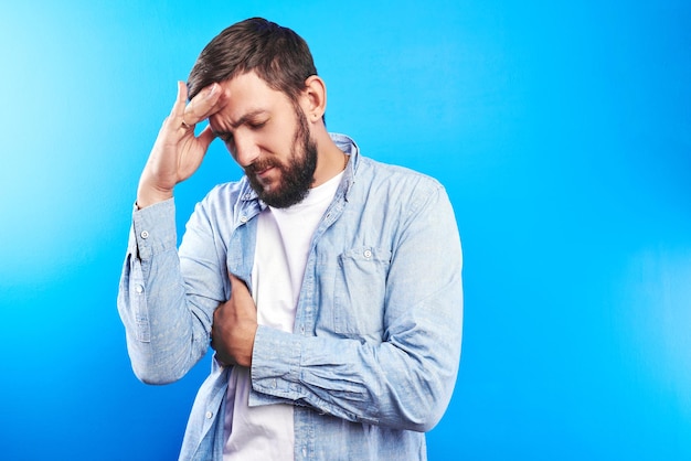 Handsome caucasian man holds head, experiencing constant stress, headache and emotional burnout at work, office worker isolated on blue background with copy space