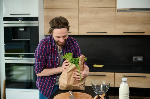 Handsome Caucasian man unpacking the paper bag with healthy food, ready for cooking delicious lunch standing by kitchen countertop in the home kitchen