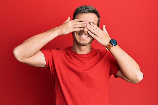 Handsome caucasian man wearing casual red tshirt covering eyes with hands smiling cheerful and funny. blind concept.