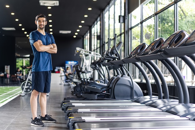 Handsome Caucasian man with beard in blue color sportswear standing and cross the arms in gym or fitness club.