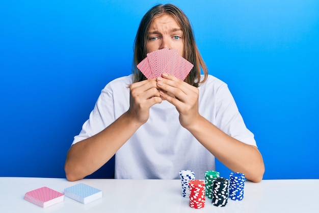 Handsome caucasian man with long hair playing gambling poker covering face with cards skeptic and nervous frowning upset because of problem negative person