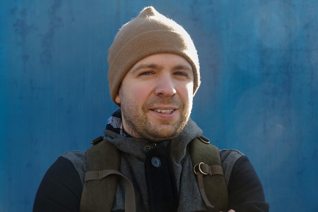 Handsome caucasian young man in hat and jacket outdoors on sunny day on blue wall