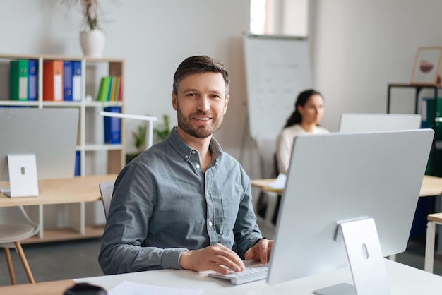 Handsome cheerful man sitting at desk using modern computer in open space office