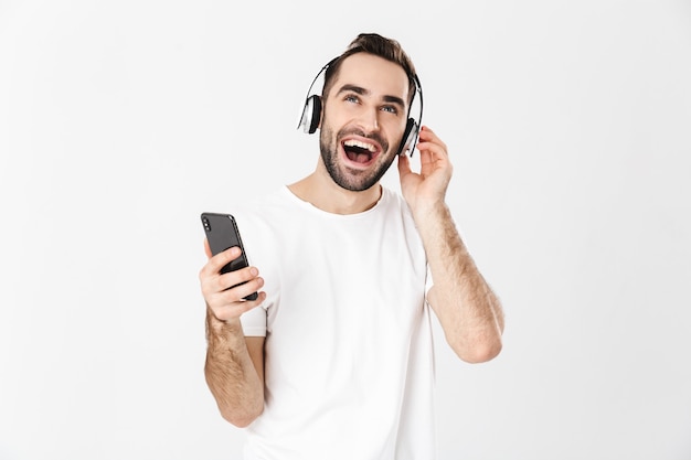 Handsome cheerful man wearing blank t-shirt standing isolated over white wall, listening to music with headphones, using mobile phone