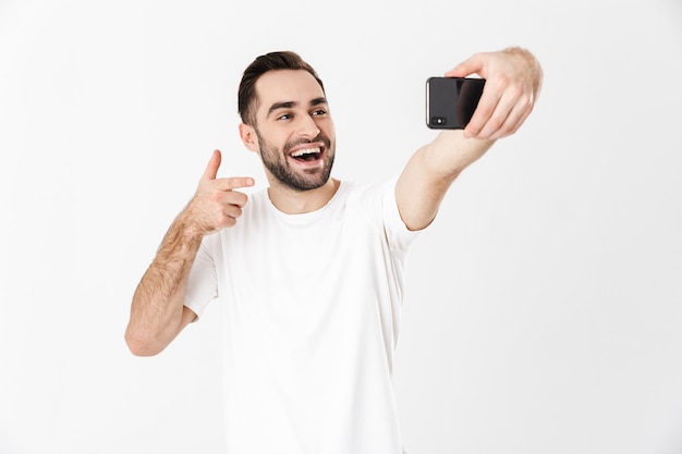 Handsome cheerful man wearing blank t-shirt standing isolated over white wall, pointing