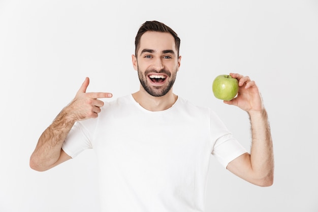 Handsome cheerful man wearing blank t-shirt standing isolated over white wall, showing green apple, pointing