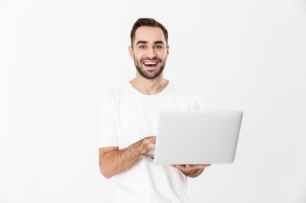 Handsome cheerful man wearing blank t-shirt standing isolated over white wall, using laptop computer