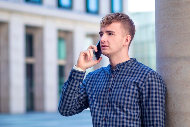 Handsome concentrated serious guy, young man in shirt talking on his cell mobile smart phone outdoors. Boy call, have conversation on smartphone. Busy businessman