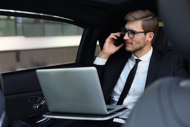 Handsome confident businessman in suit talking on smart phone and working using laptop while sitting in the car.