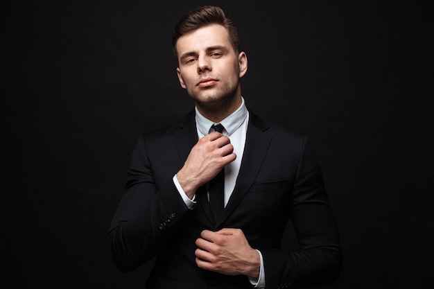 Handsome confident businessman wearing suit standing isolated over black wall, posing