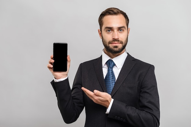 Handsome confident businessman wearing suit standing isolated over gray wall, showing blank screen mobile phone