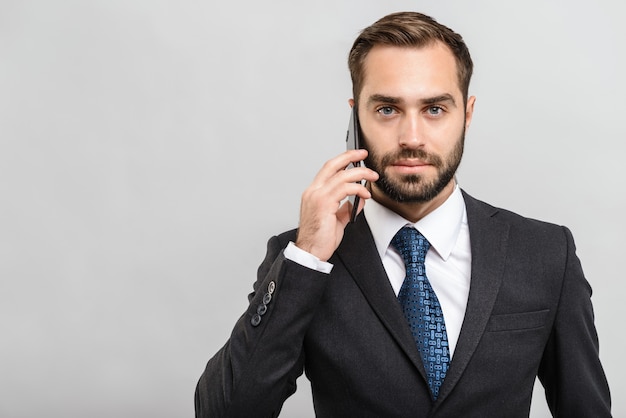 Handsome confident businessman wearing suit standing isolated over gray wall, talking on mobile phone