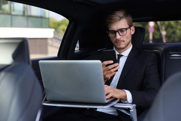 Handsome confident man in full suit looking at his smart phone while sitting in the car and using laptop.