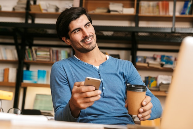 Handsome confident young man sitting at the library desk, working/studying, using laptop computer, holding mobile phone, drinking coffee