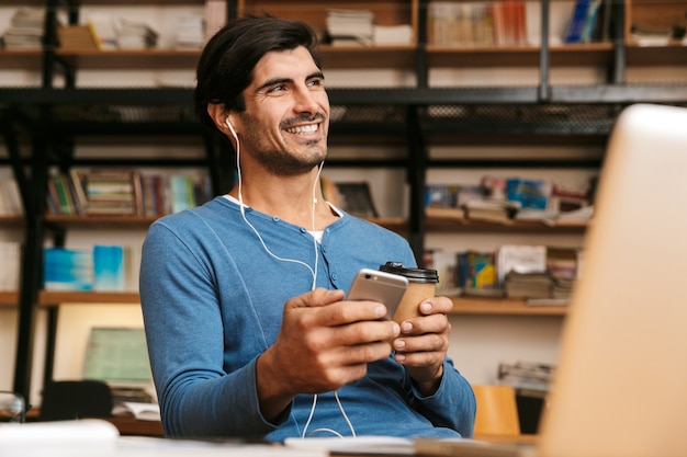 Handsome confident young man wearing earphones sitting at the library desk, working/studying, holding mobile phone