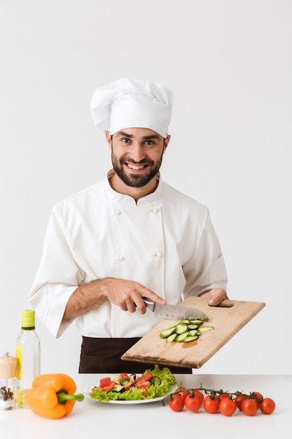 handsome cook man in uniform smiling and cutting vegetable salad on wooden board isolated over white wall