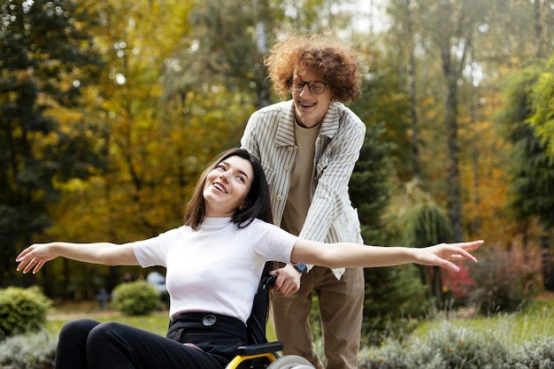 Handsome curlyhaired redhaired man carries a girl in a wheelchair Happy girl makes airplane wing gesture arms out outdoors