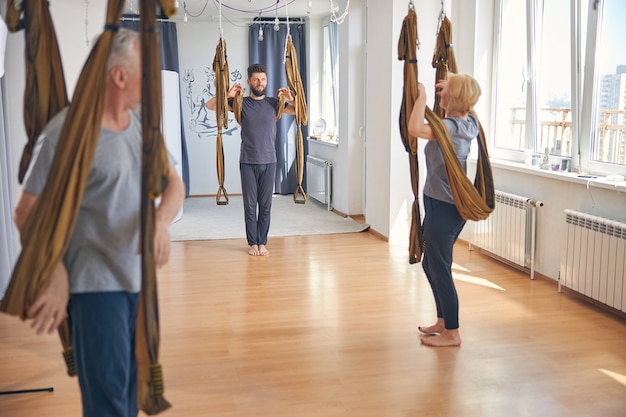 Handsome dark-haired bearded professional young Caucasian coach instructing beginner yogis before the aerial yoga class