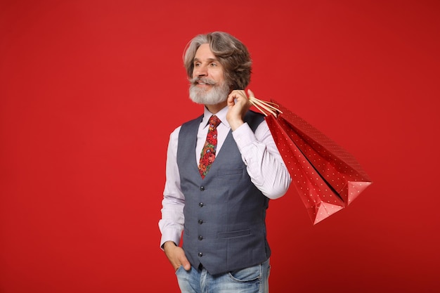 Handsome elderly gray-haired mustache bearded man in shirt vest tie posing isolated on red background. People lifestyle concept. Mock up copy space. Holding package bag with purchases after shopping.