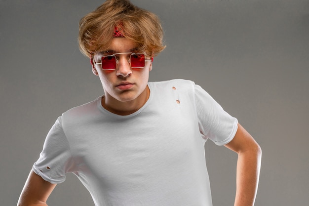 Photo handsome emotional teenager boy posing in studio against gray, guy in a gray t-shirt and red sunglasses looking at the camera