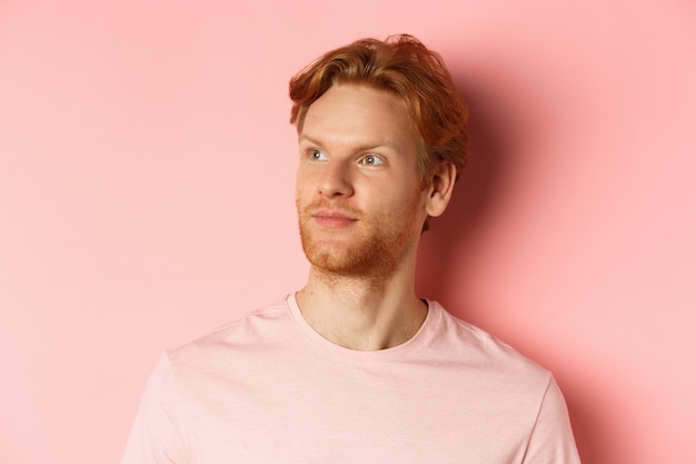 Handsome european male model with red hair and beard, turn head and looking pleased at copy space on left side, standing over pink background.