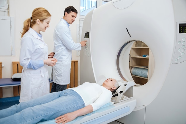 Handsome experienced male doctor looking at his patient and pressing the button while using CT scanner