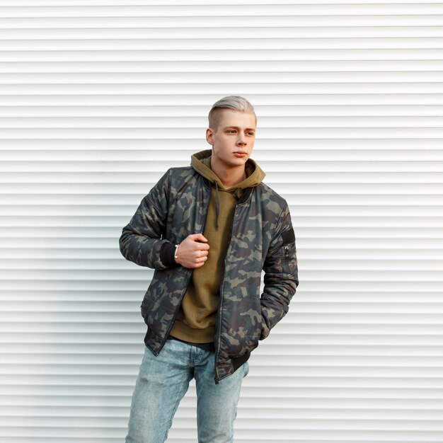 Photo handsome fashionable man in trendy military jacket with hoodie posing near white metal wall