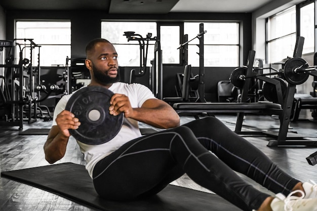 Handsome fit african man in sportwear doing crunches with damblebell in his hands at gym