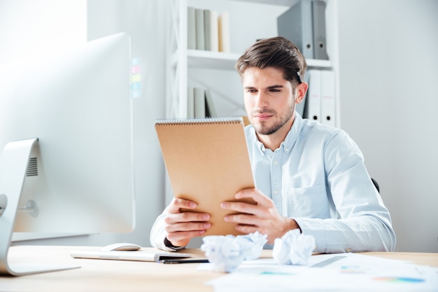 Handsome focused young man sitting and reading notes in notebook at workplace