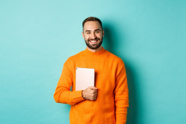 Handsome freelancer man holding laptop and smiling, standing happy over light blue background.