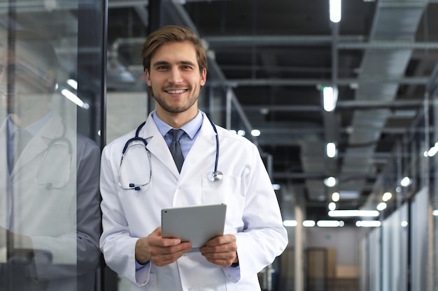 Handsome friendly young doctor on hospital corridor looking at camera, smiling.