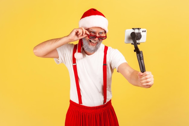 Handsome gray bearded man in santa claus costume holding smartphone on stick posing to make photo or record video, blogging. Indoor studio shot isolated on yellow background