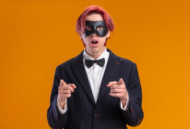 Handsome groom in suit wearing bow tie and masquerade mask  surprised pointing with index fingers  standing over orange wall