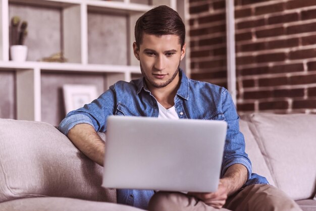 Handsome guy in casual clothes is using a laptop and smiling while sitting on couch at home