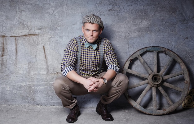 Photo handsome guy in checkered shirt with blue bow tie posing near gre concrete wall with wooden wheel.