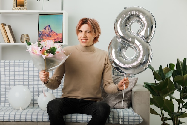 Handsome guy on happy women day holding number eight balloon and bouquet sitting on sofa in living room