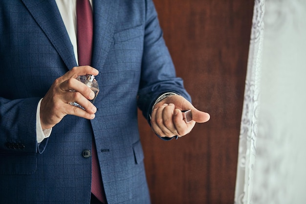 Handsome guy is choosing perfumes Elegant man in suit using colognegroom getting ready in the morning before wedding ceremony