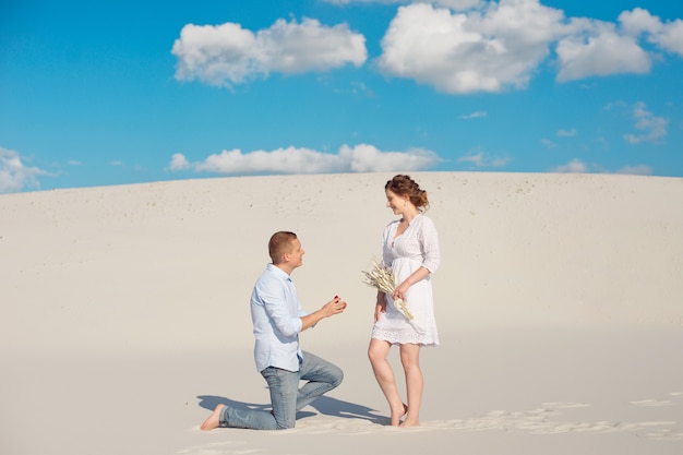 Handsome guy makes the girl a proposal for marriage, bending his knee, standing on the sand in the desert.