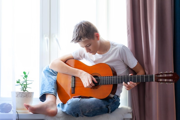 Handsome guy playing acoustic guitar while sitting on windowsill at home in his room. Boy making music playing the acoustic guitar