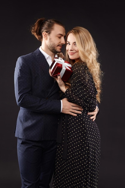 A handsome guy in a stylish suit giving a red gift box to his charming girlfriend wearing a trendy d...