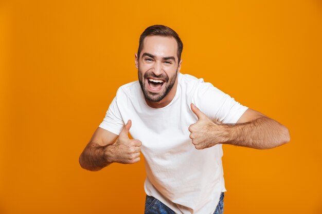 Photo handsome guy in t-shirt rejoicing and showing thumbs up while standing, isolated on yellow