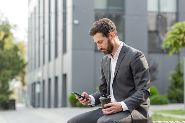 Handsome happy bearded businessman sitting on bench with cup coffee use mobile phone browse smartphone in hands reads good news and joyful smile. Outside a modern urban street background. City park