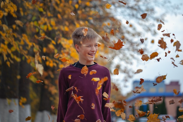 Handsome happy boy throwing the fallen leaves up, playing in the autumn park.