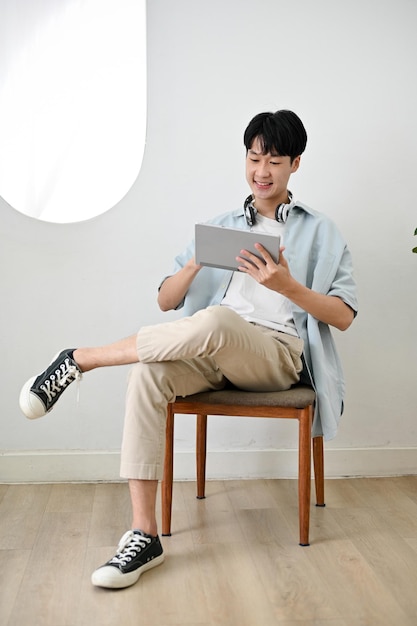 Handsome and happy young Asian man using digital tablet on a chair in minimal living room