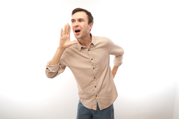 Photo handsome happy young caucasian male shouting and cupping hands around mouth isolated on white studio background. excited news attention!