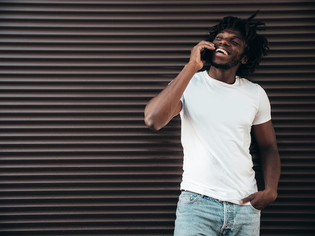 Handsome hipster modelUnshaven African man dressed in white summer tshirtFashion male with dreadlocks hairstyle Posing in the streetUsing has smartphone Talking with friends at cellphone