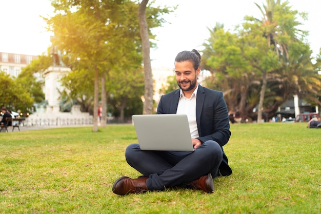 Handsome hispanic male businessman sitting lotus position in public park use laptop