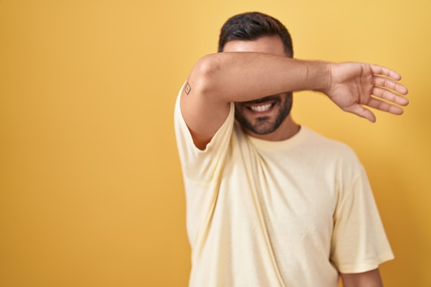 Handsome hispanic man standing over yellow background covering eyes with arm smiling cheerful and funny. blind concept.